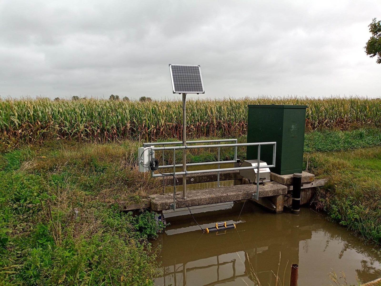 Peilstation bij een stuw met zonnepaneel tussen maisvelden, robuust buitenterrein